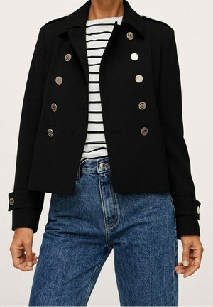 Schwarze doppelreihige Jacke mit goldenen Knöpfen über einem weiß-schwarz gestreiften Hemd, kombiniert mit hoch taillierten blauen Jeans.