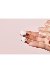 Substance crémeuse blanche en deux petites nouilles, tenue entre des doigts sur un fond rose, mettant en valeur la texture lisse et la consistance.