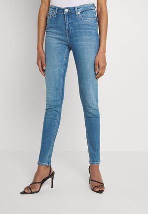 Vrouw die lichtblauwe skinny jeans en zwarte hoge hakken sandalen draagt, staat tegen een effen achtergrond.