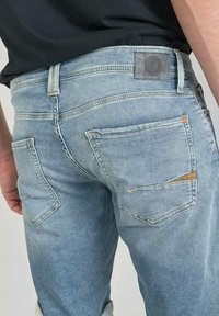 Jeans en denim bleu clair avec une finition délavée, cinq poches et une pièce sombre à la taille. Présente des coutures visibles et un design de poche distinct.