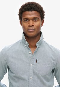 Lichtblauw, knoopshirt met kraag, zak op de linkerborst en een gestructureerde stof. Witte knopen en een klein merketiket als accent.