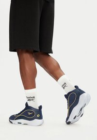 Modré športové tenisky so zlatými akcentmi, gumovou podrážkou a textúrovaným materiálom. Biely logo Reebok na ponožkách dopĺňa čierne šortky.