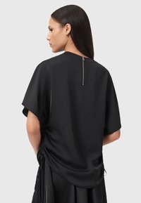 Blusa nera con maniche corte, texture liscia, dettaglio arricciato ai lati e cerniera posteriore. Design semplice e minimalista.