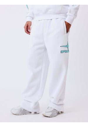 Personne portant un pantalon de survêtement blanc avec un logo bleu clair sur la cuisse, des baskets blanches et un sweat-shirt blanc, une main dans la poche.