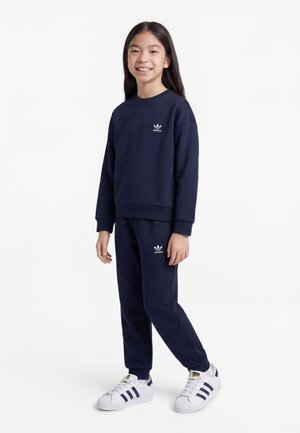 Šypsosi stovintis vaikas, dėvintis tamsiai mėlyną Adidas džemperį ir kelnes bei baltus Adidas sportbačius, stovintis priešais paprastą baltą foną.