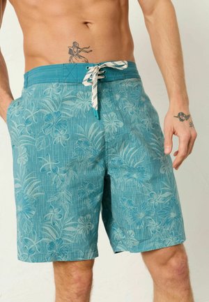 Mann trägt türkisfarbene Badeshorts mit Blumenmuster und weißem Kordelzug, zeigt Oberkörper sowie Tattoos am Bauch und an der Hand.