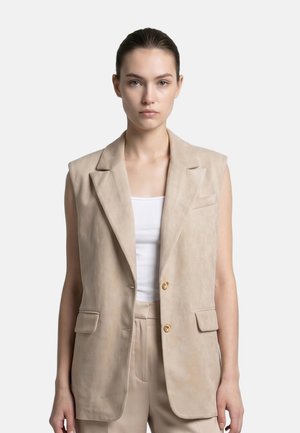 Donna con capelli scuri legati indossa un blazer beige senza maniche sopra una maglietta bianca e pantaloni beige, guarda in avanti con espressione neutra.