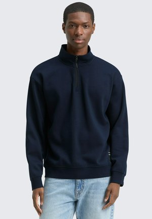 Marineblauwe kwart-zip sweatshirt in een gestructureerde stof, met ribgebreide manchetten en een relaxed fit, gecombineerd met lichte denim jeans.