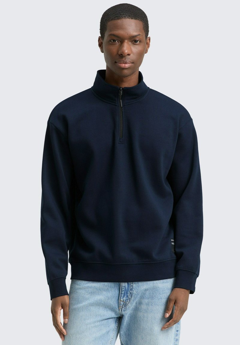 Sweatshirt bleu marine à fermeture éclair quart, en tissu texturé, avec des poignets ribbed et une coupe décontractée, associé à un jean en denim clair.