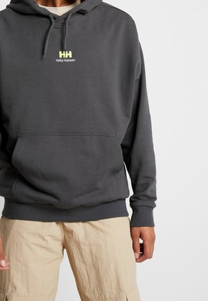 Osoba ubrana w ciemnoszarą bluzę z kapturem Helly Hansen z przednią kieszenią i ściągaczem w kapturze, w połączeniu z beżowymi spodniami typu cargo.