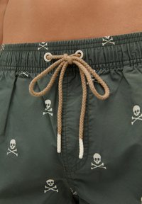 Costume de baie verde cu un model alb de cranii și cranii încrucișate. Are un brâu elastic cu un șnur bej și gauri metalice.