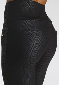 Leggings neri con motivo a spina di pesce, vita alta, texture liscia, dotati di una piccola tasca e un dettaglio in oro.