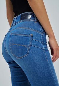 Los pantalones vaqueros de mezclilla azul presentan un diseño ajustado con una cintura de tiro medio, bolsillos traseros con costuras en crema y una etiqueta negra en la cintura.