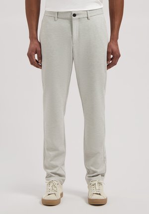 Mann trägt hellgraue Slim-Fit-Hose und weiße Sneakers, steht vor einfarbigem Hintergrund.