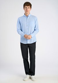 Lichtblauwe gestreepte overhemden met een borstzak, gecombineerd met zwarte broek en witte sneakers, staand tegen een effen achtergrond.