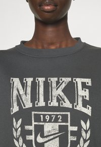 Mörkgrå sweatshirt med ribbad rund halsringning, med stor vit "NIKE"-text och en logotyp med året 1972 samt dekorativa lagerkransaccenter.