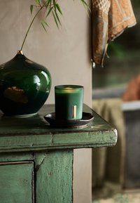 Zielona, żeberkowana świeca w szklanym świeczniku z złotym akcentem, umieszczona na ciemnej ceramicznej misie, na postarzałym zielonym drewnianym stole.