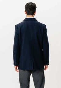 Mand står med ryggen til kameraet iført en mørk marineblå blazer og grå bukser foran en plain hvid baggrund.