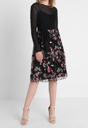 Femme portant une robe noire avec des manches longues transparentes et une jupe à motif floral, associée à des sandales à talons hauts beige, debout sur un sol blanc.