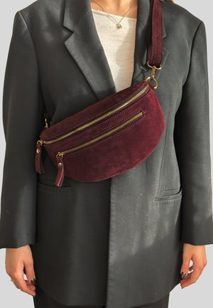 Marsupio in camoscio bordeaux con due scomparti con cerniera dorata e nappine decorative, indossato a tracolla sopra un blazer nero.