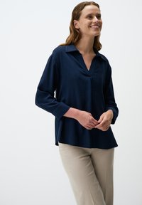 Camicia a maniche lunghe blu navy con scollatura a V e vestibilità morbida. Parte posteriore leggermente più lunga e tessuto morbido e liscio. Abbinata a pantaloni beige chiaro.