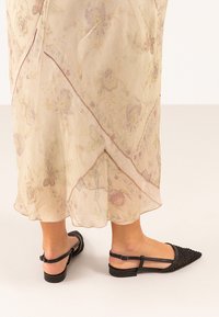 Sandalias planas negras con un diseño trenzado y correa ajustable en el tobillo; combinadas con un vestido ligero de estampado floral, que presenta un tejido suave y translúcido.