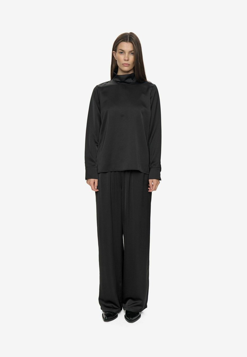Femme debout, faisant face, portant un haut noir à col roulé ample et un pantalon noir à jambes larges avec des chaussures noires à talons pointus sur fond blanc.