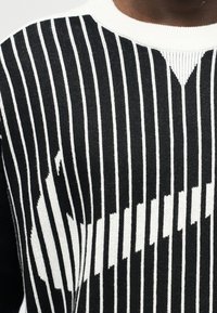 Πλεκτό πουλόβερ σε μαύρο και άσπρο με κάθετες ρίγες και ένα βαθμιδωτό, ψηφιακό λευκό μοτίβο στο στήθος και στο μπράτσο.