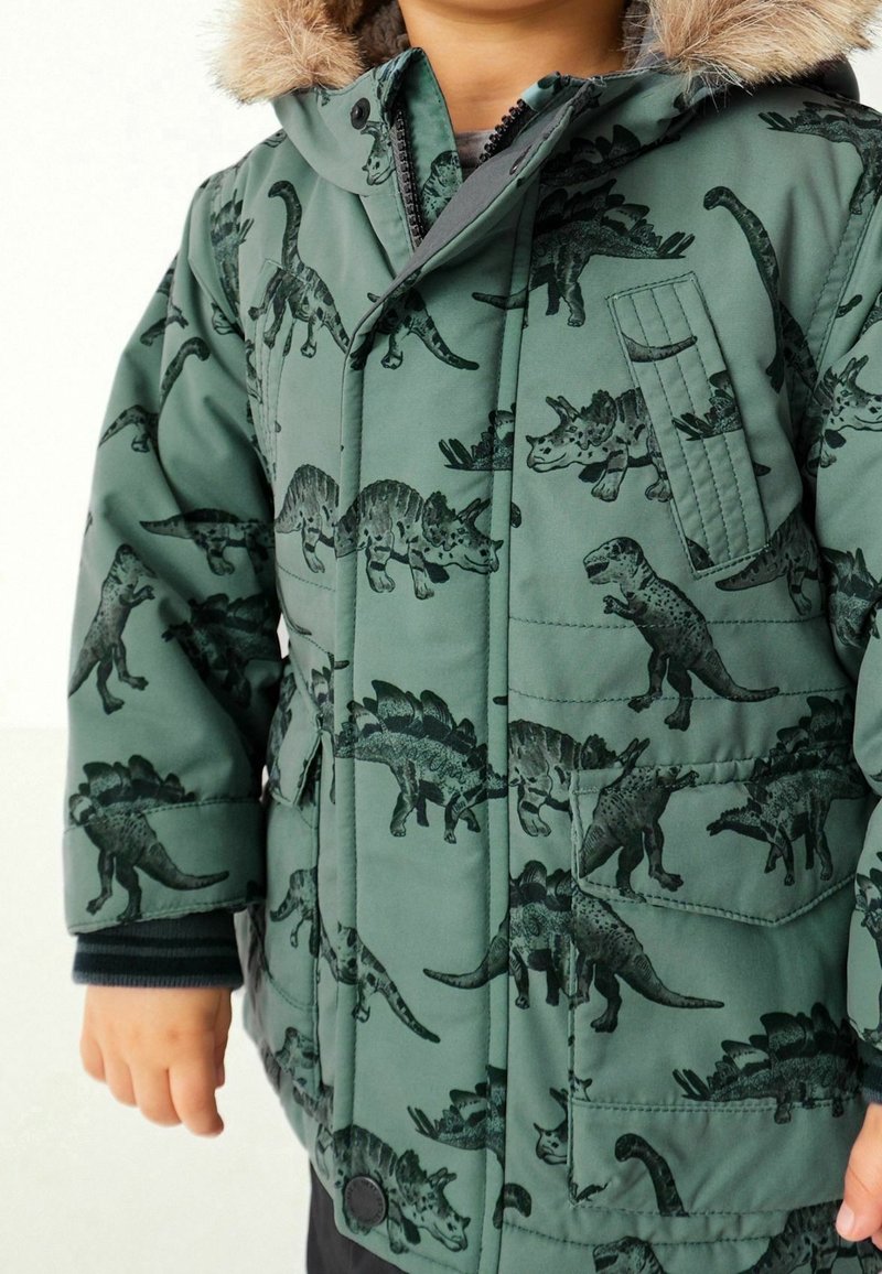 Veste matelassée verte avec un motif de dinosaures dans des tons sombres, dotée d'une capuche amovible en fausse fourrure et d'une poche poitrine. Surface du tissu texturée.