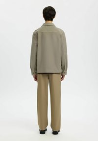 Persoon met de rug naar de camera, gekleed in een lichtgrijze jas, beige wijde broek en zwarte schoenen, op een witte achtergrond.