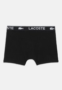 Juodos bokserio trumpikes su prekės ženklo elastinga juosta, kurioje yra balti "Lacoste" užrašai ir krokodilo logotipai.