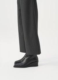Črne klinaste gležnjarje iz gladkega usnja, z ostrim nosom in dekorativnim čipkastim detajlom spredaj. V kombinaciji s sivimi hlačami.