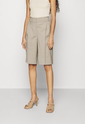 Vrouw die beige, hoog getailleerde, knielange, op maat gemaakte shorts en beige open teen haksandalen draagt, staand tegen een effen witte achtergrond.
