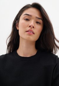 Maglione nero con scollo rotondo, dal tocco morbido, posato su uno sfondo bianco semplice. Il volto del modello è illuminato delicatamente.