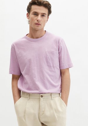 Giovane uomo con capelli ricci che indossa una camicia a maniche corte viola chiaro con taschino sul petto e pantaloni beige con le mani in tasca.