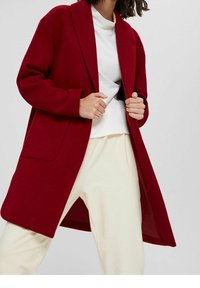Bordo płaszcz z mieszanki wełny o luźnym kroju, z otwartym przodem i dwoma przednimi kieszeniami. Noszony na białej bluzce i kremowych spodniach. Miękka tekstura.