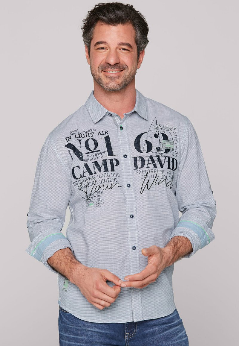 Camisa abotonada de color azul claro con texto y patrones impresos en negro, mangas largas remangadas en los puños, acentos de múltiples tonos y un cuello puntiagudo.