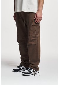 Pantalon cargo marron avec plusieurs poches, cordons de serrage à l'ourlet, associé à des baskets noires et blanches. Le matériau semble robuste et texturé.