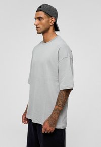 Lichtgrijze oversized t-shirt van katoen, ronde hals, korte mouwen, met een gladde textuur. Draagt met een donkere pet en losse donkere broek.