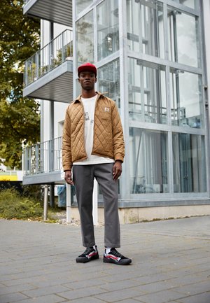 Chaqueta acolchada marrón, camiseta gráfica blanca, pantalones grises ajustados y zapatos negros con acentos rojos, frente a un edificio moderno de cristal.
