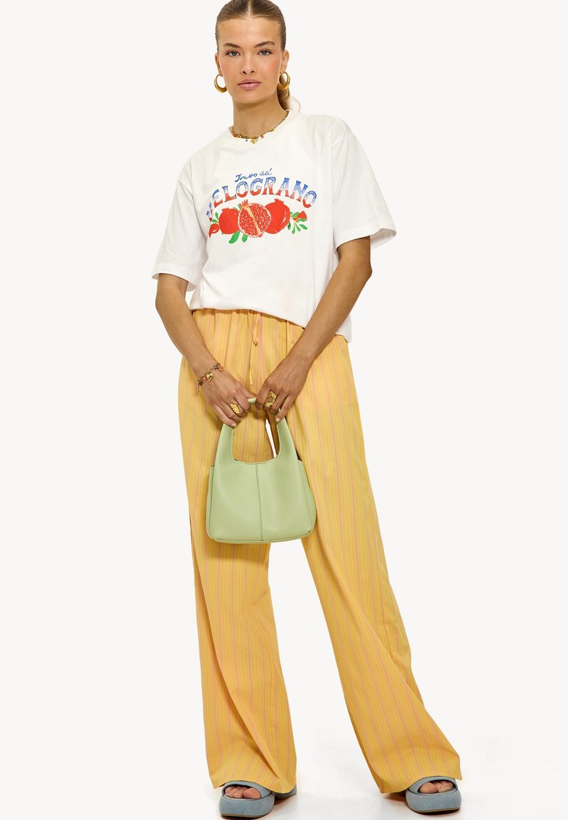 Witte grafische t-shirt met een granaatappelontwerp, gecombineerd met gele gestreepte wijde broek. Houdt een kleine groene handtas vast en draagt blauwe slippers.