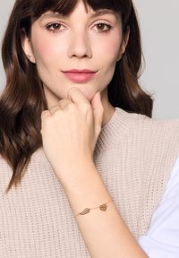 Złota bransoletka z zawieszkami w kształcie skrzydeł i serca. Design jest delikatny, z misternymi wzorami, noszona na jasnej ręce.