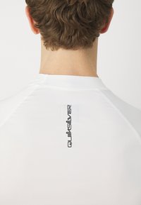 Hvit treningstrøye med en svart "Quiksilver"-logo trykket vertikalt på ryggen. Glatt tekstur med en tettsittende passform og raglanermer.