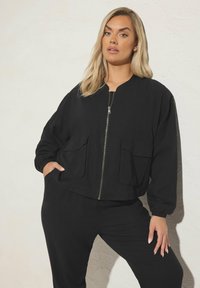 Svart bomberjacka i vävt tyg, med dragkedja fram, två stora fickor och elastiska muddar, i kombination med matchande svarta byxor.