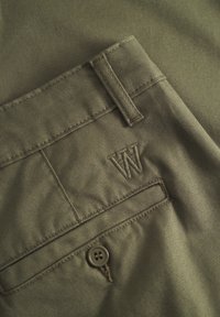 Olivgrüne Hose aus strukturiertem Stoff, mit einem Knopfverschluss und einem gestickten "W"-Logo nahe der Gesäßtasche.