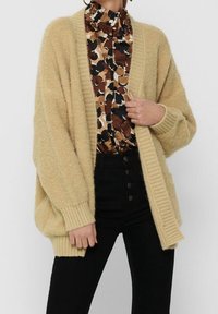 Béžový oversized kardigan s žebrovanými okraji, vyrobený z texturované příze. Nošený přes vzorované tričko s přírodními odstíny a vysokým pasem černých kalhot.