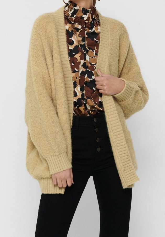 Béžový oversized kardigan s žebrovanými okraji, vyrobený z texturované příze. Nošený přes vzorované tričko s přírodními odstíny a vysokým pasem černých kalhot.