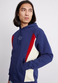 Hoodie bleu marine avec panneaux latéraux rouges et crème, poche avant, capuche avec cordon de serrage et logo sur la poitrine. Tissu doux avec une coupe décontractée.