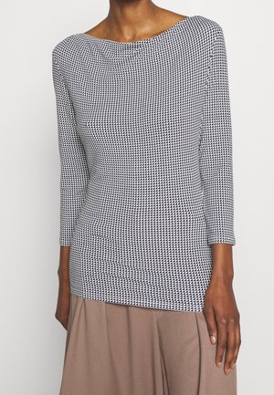 Nainen pukeutuneena pitkähihainen istuvaan yläosaan, jossa on mustavalkoinen pieni kolmiokuvio, ja beiget leveälahkeiset housut.