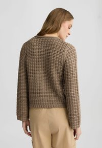 Brauner Strickpullover mit strukturiertem Muster, cropped Design und langen Ärmeln. Über beigen Hosen getragen, was einen lässigen Stil unterstreicht.